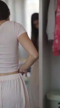 Genç bir kadın yatak odasındaki aynaya bakarken çok sayıda şık kıyafet denerken derin bir şekilde yansıtır.