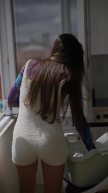 Genç bir kadın güneşli dairesinde renkli çamaşırlarını asıyor, günlük yaşamını ve evcimenliğini güzelce sergiliyor.