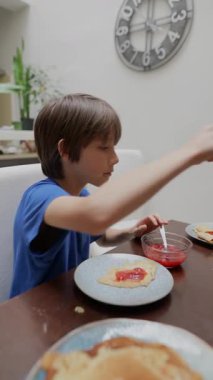 Genç, neşeli bir çocuk kahvaltıda leziz kreplerinin üstüne renkli jöle sürmekten zevk alır.