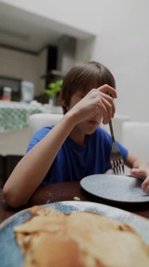 Bir çocuk mutlu bir şekilde masada oturup kreplerin tadını çıkararak sıcak ve samimi bir kahvaltı anını birlikte gösterir.