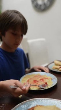 Genç bir çocuk kahvaltı sırasında sıcak bir yemek masasında çeşitli malzemelerle lezzetli kreplerin tadını çıkarıyor.