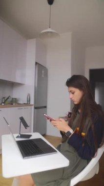 Genç bir kadın şık bir mutfak atölyesinde şık bir dizüstü bilgisayarın bulunduğu akıllı telefonuna derinden odaklanır.