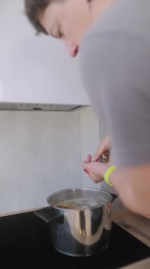 Bir insan, modern bir mutfakta yemek pişirirken fırının üzerindeki büyük bir tencereye taze malzemeler ekliyor.