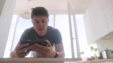 Akıllı telefon cihazıyla yakından ilgilenen, etkileşimli ve parlak bir mutfak düzenine dalmış odaklanmış genç bir adam.