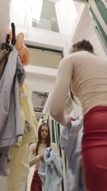 Bir kadın, çeşitli kıyafet ve kıyafetlerle çevrili bir soyunma odasında birkaç kıyafet deniyor.