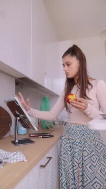 Bir kadın, elinde bir elma tutarken şık ve renkli mutfağında lezzetli tarifleri zevkle keşfeder.