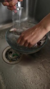 Düzenli bir mutfakta, bir insan lavaboda akan suyun altında temiz bir cam kasede taze balığı dikkatlice duruluyor.