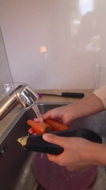 Mutfakta, bir insan lavabodaki akan suyun altında taze havuç ve canlı patlıcan yıkıyor.