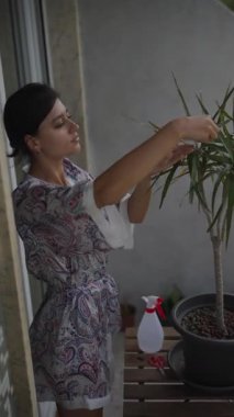 Genç bir kadın, balkonundaki manzaranın tadını çıkarırken dikkatli ve düşünceli bir şekilde ev bitkisine bakıyor.