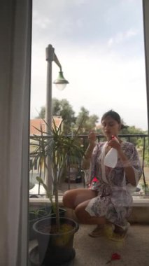 Bir kadın, güzel bir manzarayı gösterirken bir balkonda bulunan gür iç mekan bitkileriyle özenle ilgilenir.