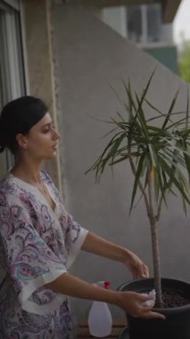 Bir kadın balkonda saksısıyla nazikçe ilgilenir, özgüven ve doğa takdirini gösterir..