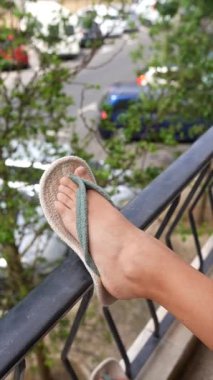 Renkli bir sandaletin içinde rahat bir şekilde duran rahat bir ayak, canlı bir caddeye bakan bir balkon korkuluğunda dinleniyor.