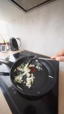 Bir insan, sıcak bir mutfaktaki indüksiyon sobasına yerleştirilmiş tavada ustalıkla doğranmış soğan karıştırıyor.