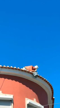 Kendini işine adamış bir işçi, güzel mavi gökyüzünün altında, pembe kıvrımlı bir çatı üzerinde gerekli bakımı yapıyor.