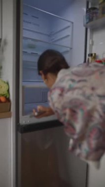 Bir kadın düşünceli bir şekilde buzdolabını araştırıyor, çeşitli atıştırmalıklar ve yemek seçenekleri düşünüyor, günlük hayatı somutlaştırıyor.