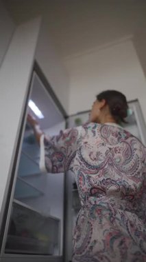 Renkli bir bornoz giymiş bir kadın buzdolabını karıştırıyor. Yemek ve atıştırmalıklar için yiyecek seçimlerini düşünüyor.