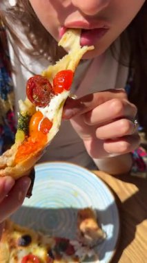 Bir insan sıcak güneşin altında, taze malzemelerle dolu canlı bir dilim pizzanın tadını çıkarıyor.