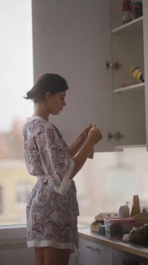 Sabahlık giymiş bir kadın mutfak dolabındaki eşyaları inceliyor, sakin bir sabah rutini sergiliyor..