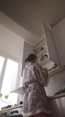 Konforlu bir bornoz giyen bir kadın mutfak dolaplarındaki çeşitli eşyalara uzanıyor ve ev düzenini etkili bir şekilde sergiliyor.