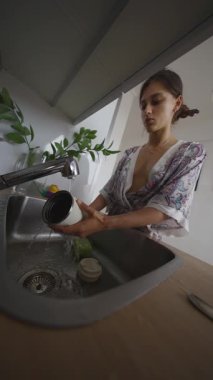 Rahat bornozlu bir kadın lavaboda mutfak malzemelerini yıkıyor, şık ve düzenli bir ev dekorasyonu gösteriyor.