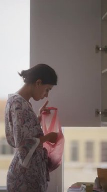 Bir kadın rahat bir mutfakta, şık desenli bir bornoz giyerken düşünceli bir şekilde pembe bir çantayı inceliyor.