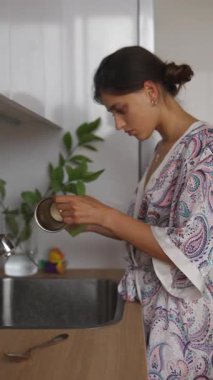 Güzel desenli bir bornoz giymiş genç bir kadın modern mutfak ortamında özenle çatal bıçak yıkıyor.