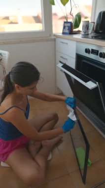 Genç bir kadın, modern mutfağındaki bir fırının cam kapısını temizliyor..