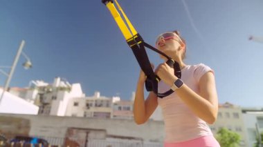 Bir kadın açık hava TRX eğitimine katılır, güneşli bir ortamda spor ve güç egzersizleri yapar..