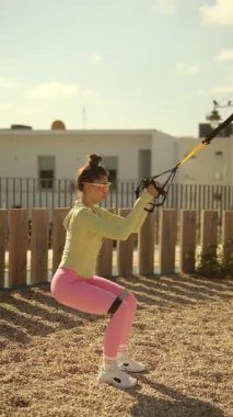 Bir kadın açık hava ortamında süspansiyon kayışları kullanarak çömelme hareketleri sergiliyor..