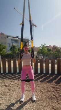 Formda bir kadın güneşli bir açık hava parkında süspansiyon egzersizi yapıyor, temiz hava ve enerjinin tadını çıkarıyor.