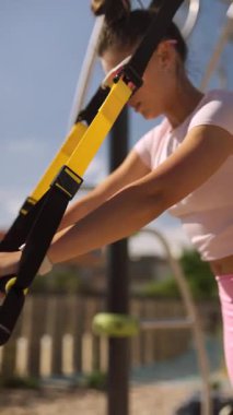 Genç bir kadın, güneşli bir ortamda yoğun bir egzersiz için TRX süspansiyon teçhizatı kullanıyor.