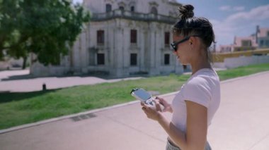 Tarz sahibi genç bir kadın güzel bir açık hava manzarasının tadını çıkarırken akıllı telefonuyla mutlu bir şekilde etkileşime girer.
