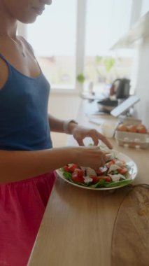 Bir kadın renkli bir salataya çeşitli taze malzemeler ekliyor. Parlak, davetkar bir mutfak ortamında.