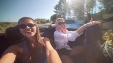 İki arkadaş mutlu bir şekilde güneşli bir yolculuğun tadını çıkarıyorlar, üstü açık bir arabada keyifli anları yakalıyorlar.