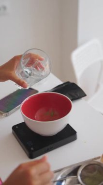 Bir insan, mutfak ölçeği cihazına yerleştirilmiş kırmızı bir kasenin içindeki çeşitli malzemeleri titizlikle tartar.