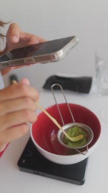 Parlak bir mutfakta yönlendirme için elek ve akıllı telefon kullanan bir birey dikkatlice matcha hazırlıyor..