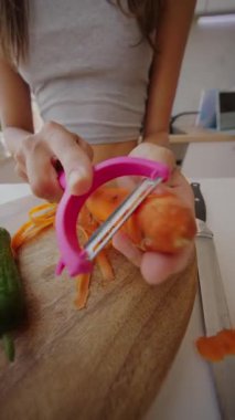 Bu resim, parlak bir mutfakta taze sebzeleri ustalıkla hazırlamak için pembe bir soyucu kullanan ellerin yakın çekimini gösteriyor.