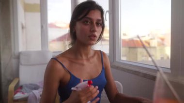 Genç bir kadın neşeyle gülümser ve serinletici bir manzarayla rahat evinde dinlenirken ferahlatıcı bir içkinin tadını çıkarır.