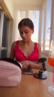 Pembe bluzlu bir kadın parlak bir kafede otururken makyaj yapıyor.