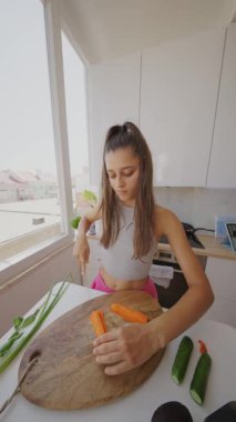 Genç bir kadın güzel bir manzara sunan parlak, havadar bir mutfakta ustalıkla taze sebzeleri doğruyor.