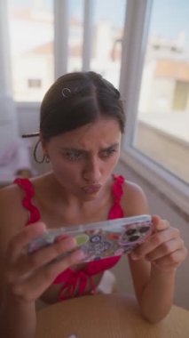 Neşeli genç bir kadın, neşeli bir şekilde akıllı telefonuyla etkileşime geçiyor, güneşli bir ortamda çeşitli duyguları ifade ediyor.