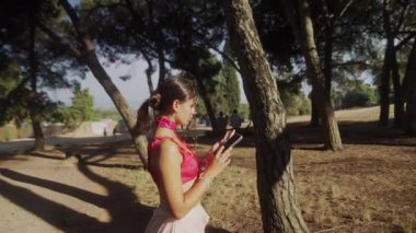 Genç bir kadın, yemyeşil ağaçlar ve doğa ile çevrili güneşli bir parkta akıllı telefonuyla mutlu bir şekilde iletişim kuruyor.