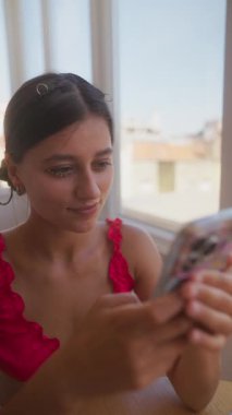 Rahat bir şekilde oturan genç bir kadın, akıllı telefonuyla derinden bağlanır, parlak, güneşli bir odada saf neşe saçar.