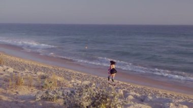 Bir kadın neşeli bir şekilde sahilde koşuyor. Renkli dalgalar canlı bir günbatımına çarpıyor.