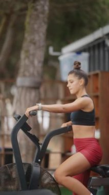 Genç bir kadın dışarıda durağan bir bisikletle egzersiz yapıyor. Forma ve sağlığa olan bağlılığını gösteriyor.
