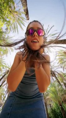 Neşeli ve neşeli genç bir kadın, şık güneş gözlükleriyle birlikte, baş döndürücü palmiye ağaçlarının altında neşe içinde poz veriyor.