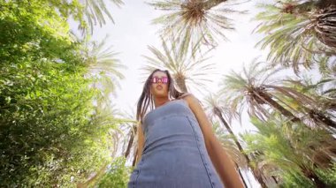 Güneşli bir günde, şık bir genç kadın, palmiye ağaçlarının arasında kot elbiseyle poz verir ve canlı bir atmosfer yaratır.