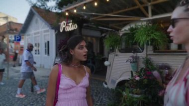 İki genç kadın yeşilliklerle dolu canlı bir kafede konuşuyor.