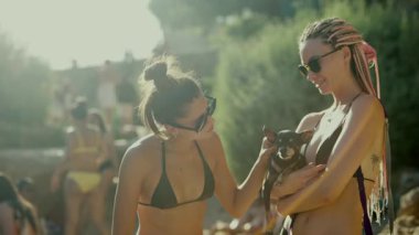 İki arkadaş, neşeli bir kumsal anının keyfini çıkarırlar. Parlak güneşin altında sevimli bir köpekle neşeyle etkileşirler.