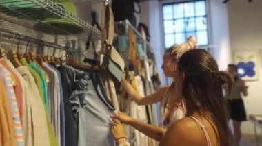 İki genç kadın zamanlarını moda bir butikte çeşitli şık kıyafet seçeneklerini araştırarak geçiriyorlar.